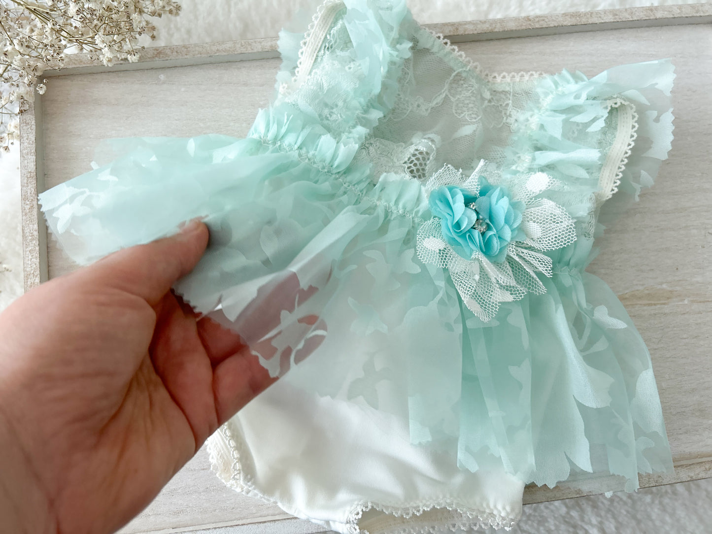 Mint green lace baby outfit with a flower headband held by a hand on a light wooden surface.