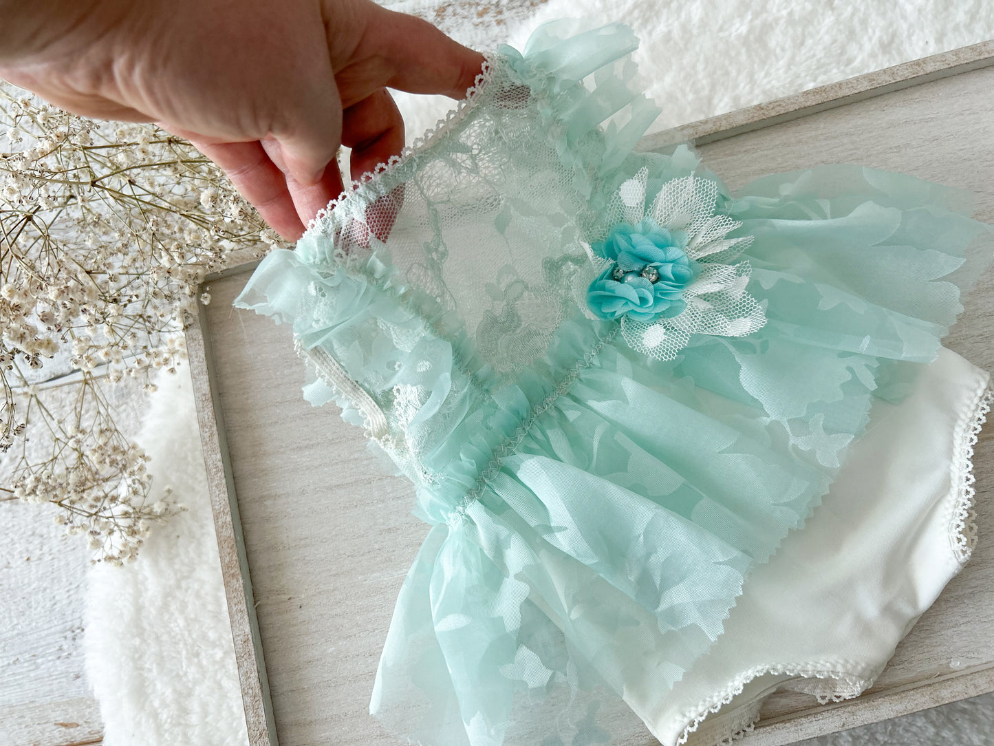 Light mint green lace newborn romper with a flower detail held by a hand on a light wooden surface.