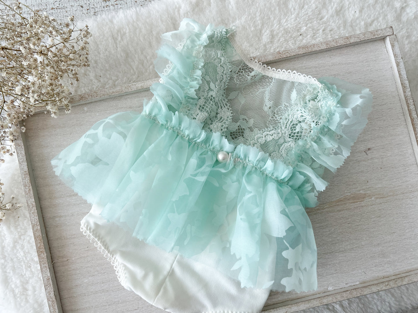 Light mint green lace baby romper with lace and ruffles on a wooden surface.