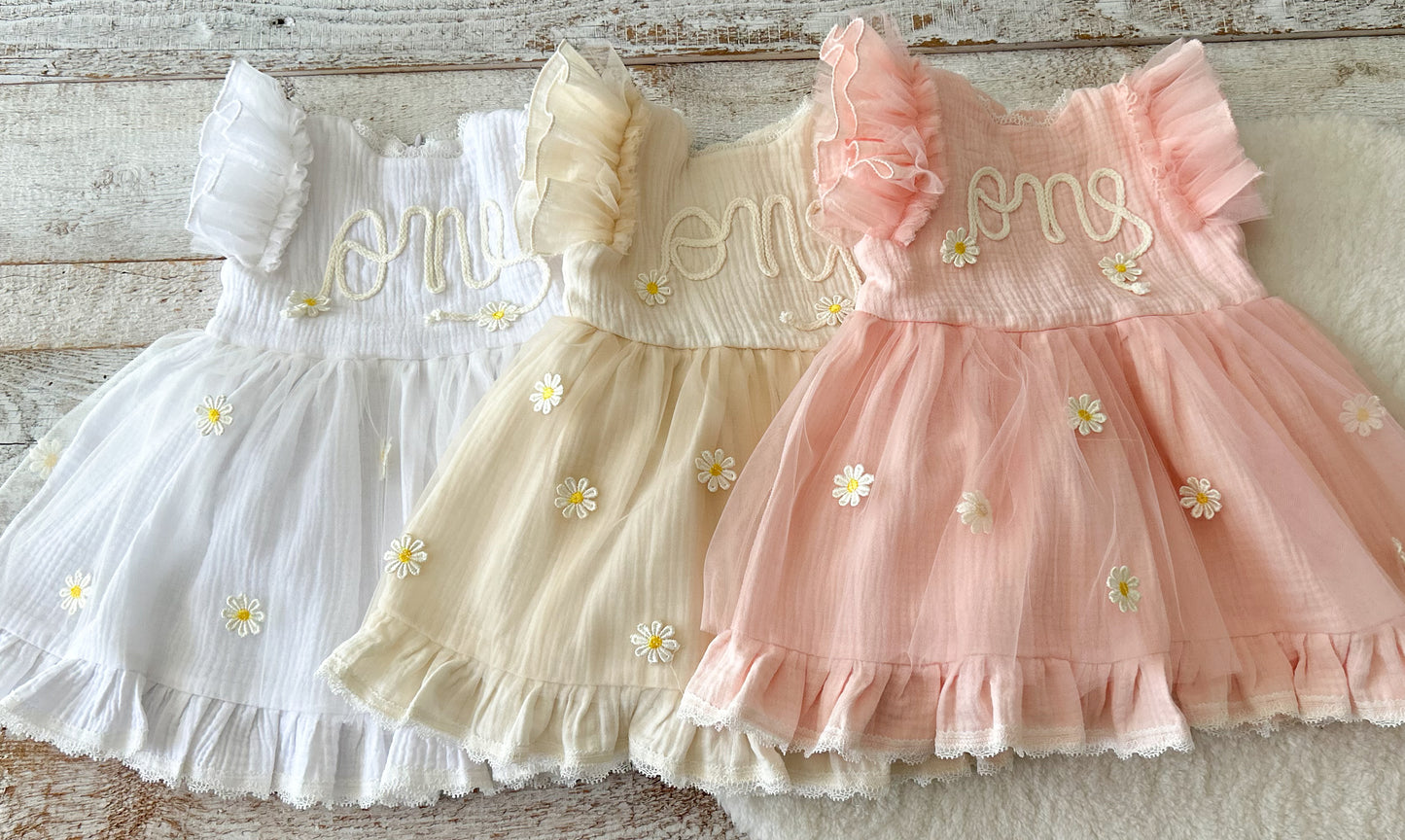 Three baby dresses in white, beige, and pink with floral embroidery on a wooden surface.