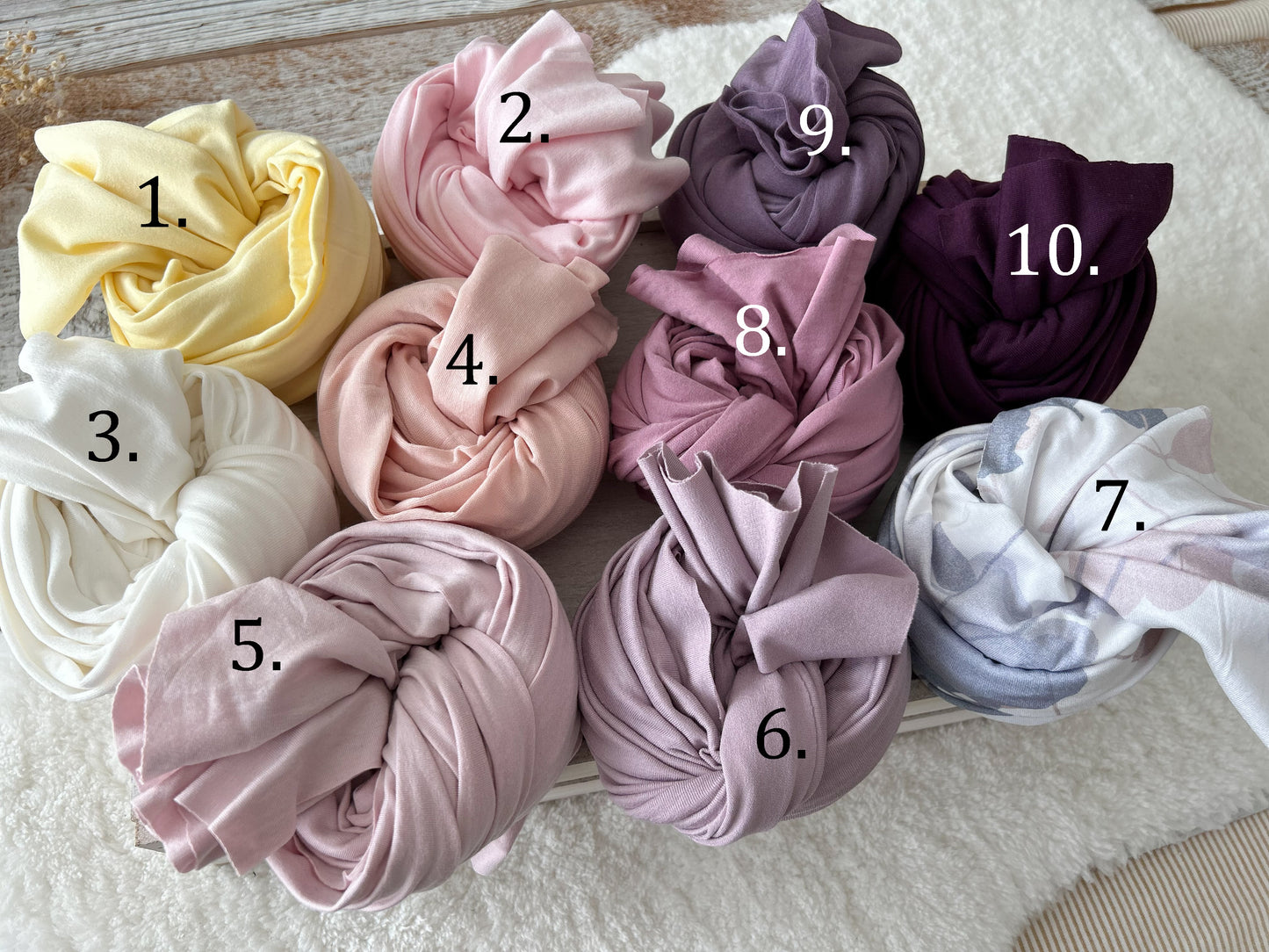 Rolled cotton swaddle wraps in 10 soft and rich tones for newborn photography, displayed on a cozy white textured surface.