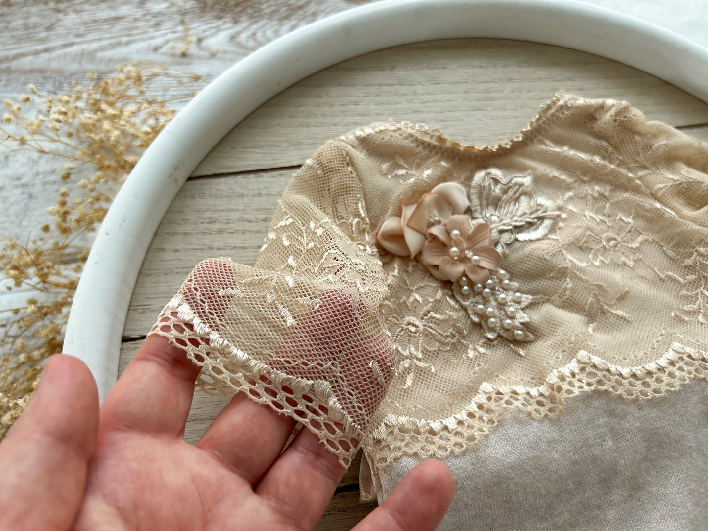 Newborn photo outfit in beige lace. Close-up of a hand holding a piece of lace fabric with floral embellishments on a wooden surface.