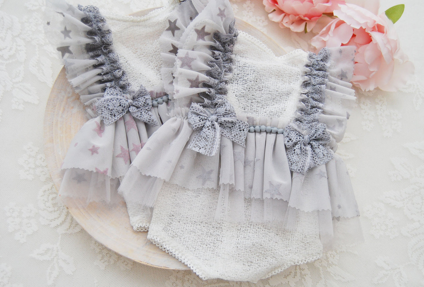 Newborn lace romper photo prop in light gray with ruffles and star pattern, displayed on a plate with a floral background and a pink flower.