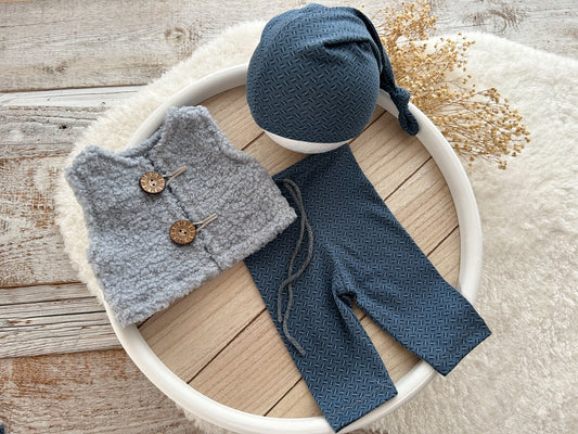 Newborn photography outfit set with blue textured pants, matching hat, and gray curly vest with wooden buttons, styled on a white tray.