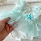 Mint green lace baby outfit with a flower headband held by a hand on a light wooden surface.