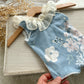 Newborn girl romper in blue fabric with floral print. It features ivory chiffon ruffles around the neckline. Ideal as newborn photo prop. 