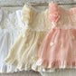 Three baby dresses in white, beige, and pink with floral embroidery on a wooden surface.