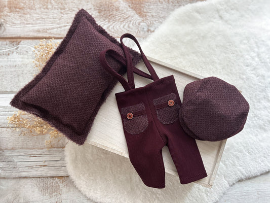 Newborn boy photo outfit in burgundy red with suspender pants, matching flat cap, and posing pillow on light wood background