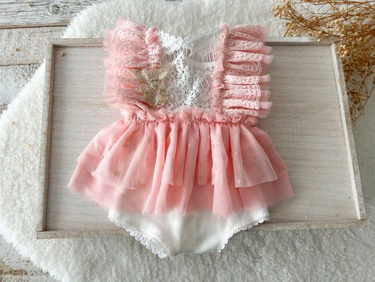 A delicate baby girl romper in candy pink with layered chiffon ruffles and white lace bodice, styled flat on a soft wooden background.