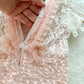 Back of pink baby romper with V-cut, pearl trim, and tied organza bows on a white textured surface.