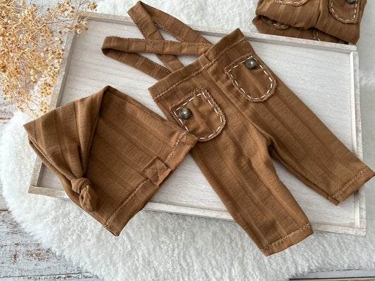 Newborn boy photo outfit with brown suspender pants and matching knot sleepy hat, laid flat on white background