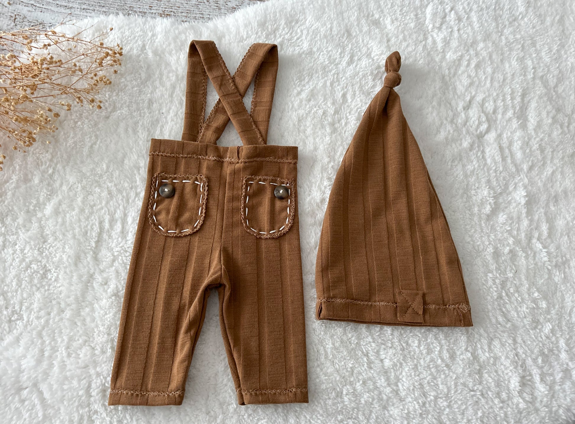 Brown newborn outfit with ribbed suspender pants and matching hat displayed on a textured cream blanket.