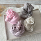Four rolled newborn photo wraps in soft cotton fabric: light pink, dusty purple-gray, light purple, and beige speckled, displayed on a wooden tray.