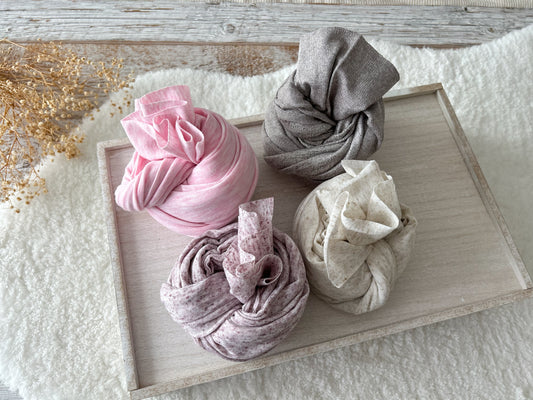 Four rolled newborn photo wraps in soft cotton fabric: light pink, dusty purple-gray, light purple, and beige speckled, displayed on a wooden tray.