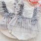 Newborn lace romper photo prop in light gray with ruffles and star pattern, displayed on a plate with a floral background and a pink flower.